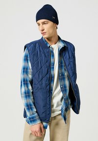 Gilet trapuntato blu navale con tasche laterali, indossato sopra una camicia a scacchi blu e bianca, abbinato a un berretto lavorato a maglia blu scuro.