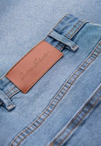 Denim kangas vaaleansinisena, jossa on tekstuurinen nahkapaikka, johon on kohokuvioitu sana "Ombre" ja kontrastoivia ompelutäytteitä.