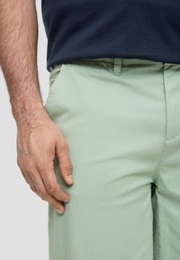 Pantalon vert clair en tissu doux, doté de poches latérales, d'une texture subtile et d'une coupe ajustée, associé à une chemise bleu foncé.