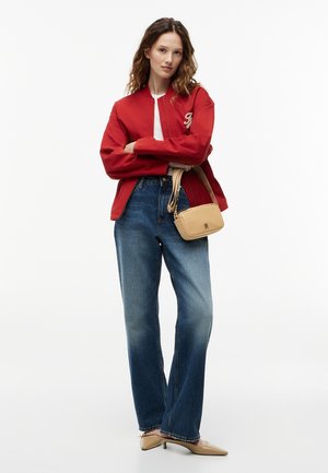 Vrouw met een rode jas, witte top, blauwe spijkerbroek, beige puntige hakken en een beige schoudertas, staand met gekruiste armen.