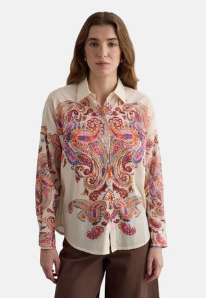 Vrouw draagt een crèmekleurig hemd met knopen met kleurrijke roze, oranje en paarse paisleypatronen en een bruine broek, kijkend naar voren.