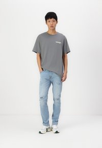 Szara bawełniana koszulka z krótkim rękawem i białym logo, zestawiona z jasnoniebieskimi, zniszczonymi dżinsami oraz białymi sneakersami z zielonymi akcentami.