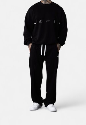 Persona con sudadera y pantalones negros con cordones blancos y zapatillas blancas, de pie con las manos en los bolsillos sobre fondo blanco.