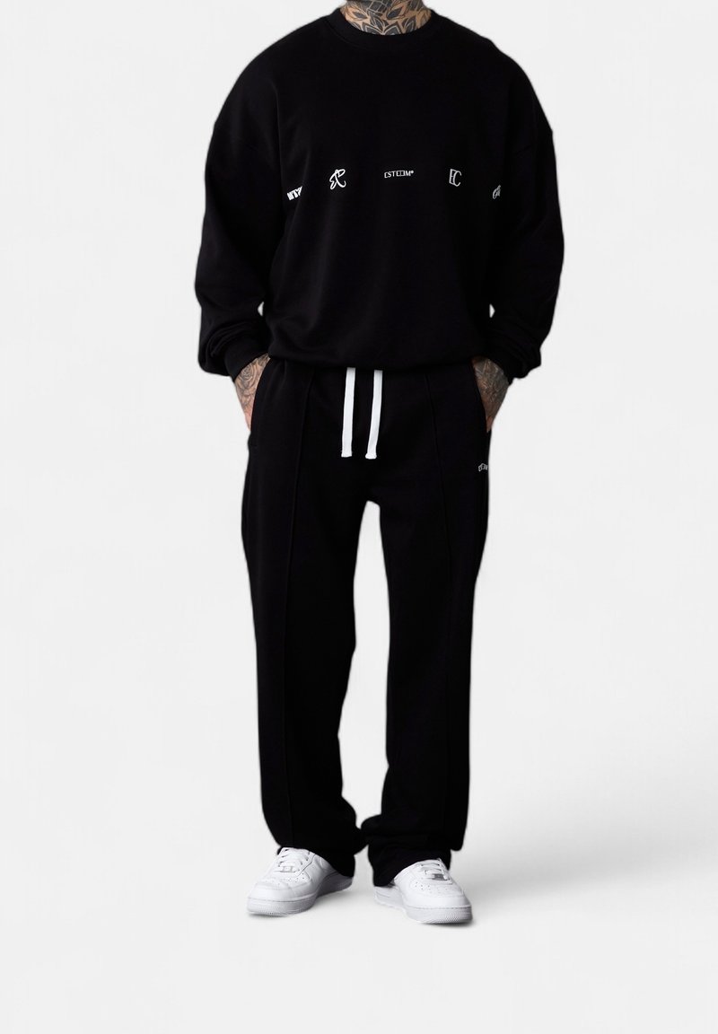 Personne portant un sweat-shirt noir et un pantalon avec des cordons blancs et des baskets blanches, debout les mains dans les poches sur fond blanc.