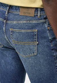 Temno modri denim kavbojke z rumenimi šivi, ki imajo usnjen znak "BOGGI" na pasu in detajl na zadnjem žepu.