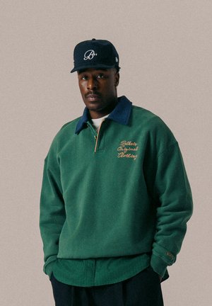 Pullover in pile verde con colletto blu, caratterizzato da testo ricamato in oro. Abbinato a un cappellino blu con logo.