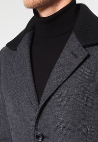 Uomo con barba che indossa un cappotto grigio scuro a trama con colletto nero sopra un maglione a collo alto nero.