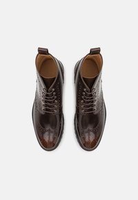 Botas de cuero marrón con cordones, detalles de brogue, acabado liso, puntera redondeada y suela oscura en contraste. Presentan una altura hasta el tobillo y costuras decorativas.
