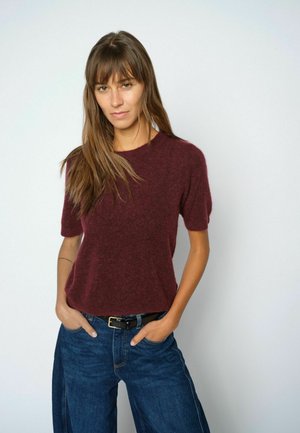 Junge Frau mit langen braunen Haaren, die einen burgunderfarbenen Kurzarmpullover und blaue Jeans trägt, mit den Händen in den Taschen, steht vor einem schlichten Hintergrund.