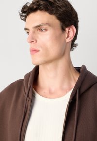 Brown zip-up hoodie with a drawstring hood, worn over a cream ribbed shirt. Smooth fabric with a relaxed fit and minimal stitching details.