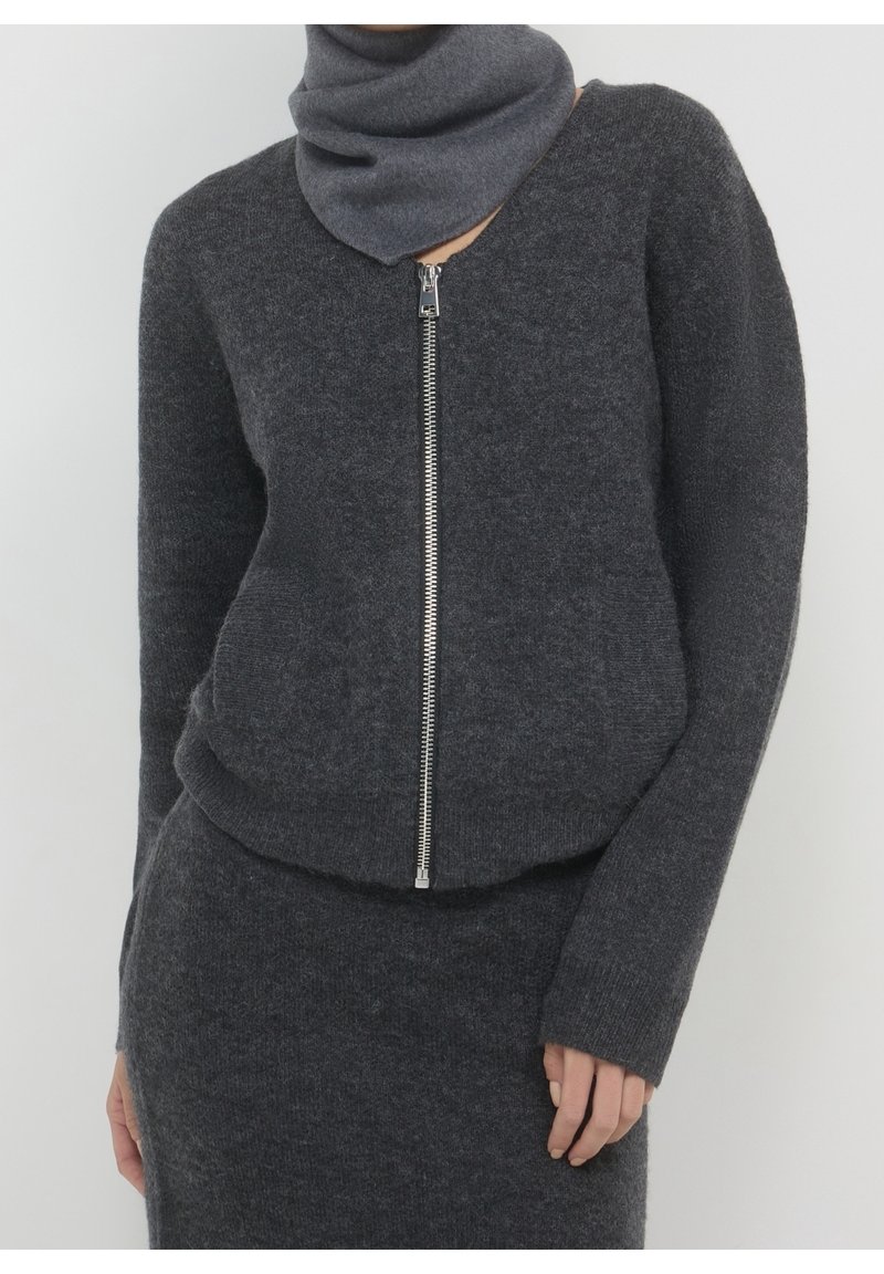 Personne portant un pull zippé gris foncé et une jupe assortie avec une écharpe grise couvrant le cou, sur un fond clair uni.