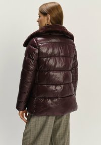 Burgunderfarbene Pufferjacke mit einem Kunstpelzkragen, ausgestattet mit gestepptem Textil und einem leicht glänzenden Finish, von hinten gezeigt.