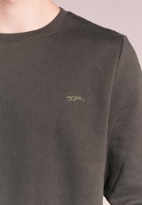 Sudadera de color verde oscuro hecha de tela suave, con un cuello redondo acanalado y un pequeño logo de tigre bordado en el lado izquierdo del pecho.
