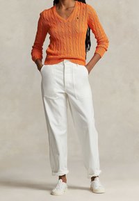 Pull orange en maille torsadée avec col en V, associé à un pantalon blanc ample et des baskets blanches. Le pull présente des poignets et un ourlet côtelés.