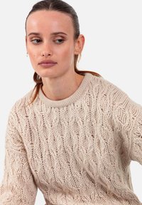 Pull en tricot beige clair présentant un motif texturé ondulé avec un col ras du cou côtelé. Tissu doux avec une coupe ample.