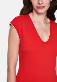 Rotes Kleid mit tiefem V-Ausschnitt, kurzen Ärmeln und weißem Besatz entlang des Ausschnitts. Der Stoff hat eine glatte, elastische Textur.