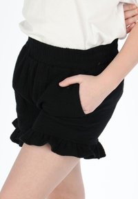 Schwarze Shorts mit einem elastischen Bund, volanten Saum und seitlichen Taschen, aus weichem Stoff, gezeigt mit einem weißen Oberteil.