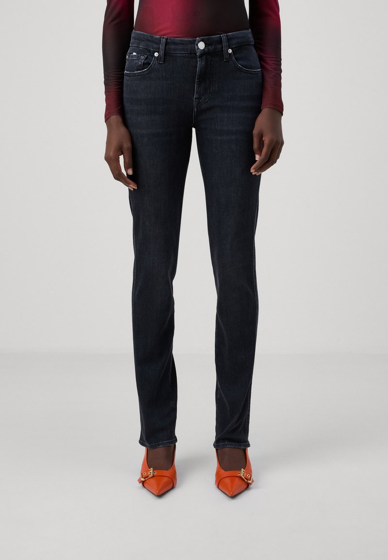 Jeans de mezclilla oscura con un diseño ajustado, que cuentan con cinco bolsillos y un cierre de botón. Combinados con tacones naranjas puntiagudos.