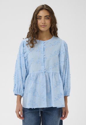 Kobieta z długimi kręconymi włosami, ubrana w jasnoniebieską haftowaną bluzkę z guzikami na przodzie i bufkami o długości 3/4, zestawioną z dżinsami.