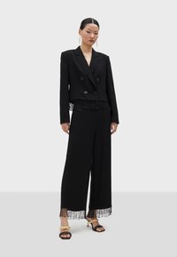 Svart kort dubbelknäppt kavaj med fransar, i kombination med vida byxor med fransdetaljer och öppna klackskor.