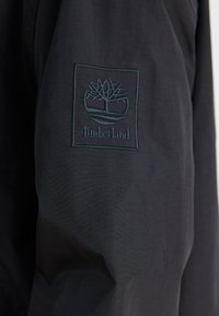 Zwarte stoffen jas mouw met een geborduurd Timberland-logo met een boomontwerp in donkergroen, omringd door een rechthoekige rand.