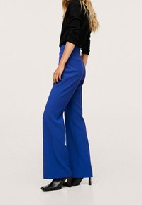 Pantalon bleu à jambes larges en tissu lisse, avec une taille haute, un pli sur le devant, porté avec des bottes noires à talons.