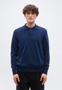 Άνδρας με σγουρά μαλλιά που φοράει μπλε σκούρο μακρυμάνικο πόλο και μαύρο παντελόνι, στέκεται μπροστά σε απλό φωτεινό φόντο.