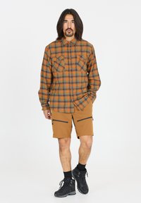 Ruudullinen paita oranssin, vihreän ja ruskean sävyissä, jossa on kaksi rinta-taskua; yhdistetty ruskeisiin shortseihin, joissa on vetoketjulliset taskut, sekä mustiin saappaisiin.