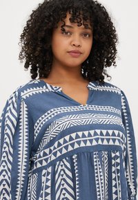 Top blu con motivo a fantasia e scollo a V. Presenta disegni geometrici bianchi e pieghe. Realizzato in un materiale leggero con maniche lunghe.