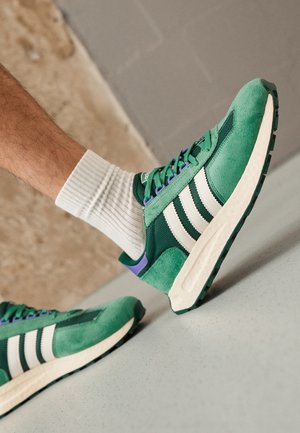 Grønne og hvide sneakers med lilla detaljer båret med hvide ribstrikkede sokker på en glat lysegrå gulv.
