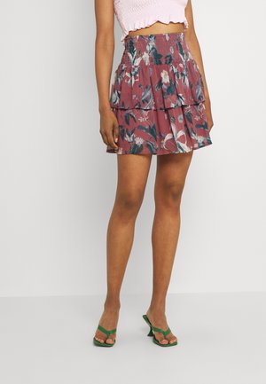 Kvinde iført en pink croppet top og en lagdelt blomsterprikket vinrød mininederdel med grønne hælede sandaler, stående mod en ensfarvet baggrund.