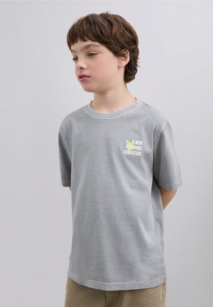 Camiseta gris de manga corta con cuello redondo, con un pequeño texto gráfico multicolor que dice "A NEW SCALPERS ADVENTURE" en el pecho.