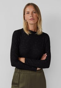 Vrouw met blond haar in een zwart longsleeve met een subtiel patroon en olijfgroene high-waisted broek, met gekruiste armen, neutrale achtergrond.