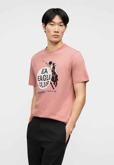 Νεαρός με μαύρα μαλλιά φοράει ροζ μπλουζάκι με γραφικό παίκτη πόλο και το κείμενο «EA Eagle Club Emporio Armani», συνδυασμένο με μαύρο παντελόνι.