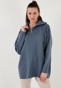 Gaiši zils džemperis ar augstu apkakli un pus-zip aizdari. Ievērojams ribota apkakles apdare un brīvs piegriezums, izgatavots no mīkstas auduma.