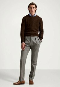 Un pull en tricot marron porté par-dessus une chemise à col bleue, associé à un pantalon gris à plis. Des chaussures en cuir marron complètent le tout.
