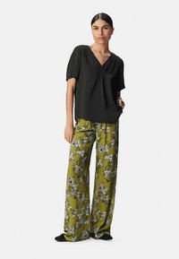 Schwarzes Kurzarmshirt mit V-Ausschnitt; lockere, weit geschnittene Hosen in Olivgrün mit floralem Muster. Flache schwarze Schuhe.