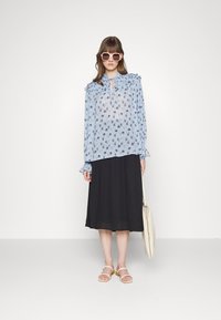 Blusa floral azul claro com ombros franzidos, saia preta plissada e sandálias bege. Modelo segura uma carteira creme e usa óculos de sol.