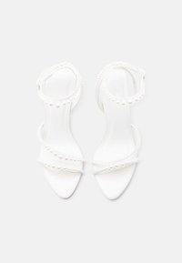 Λευκά πέδιλα με ανοιχτά δάχτυλα, λουριά στον αστράγαλο και διακοσμητικά μαργαριταρένια στολίδια κατά μήκος των λουριών και των ακμών, τοποθετημένα σε απλό λευκό φόντο.