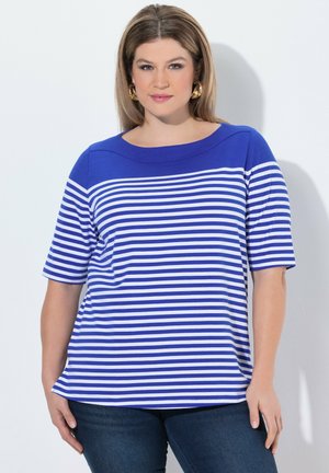 Frau trägt ein blau-weiß gestreiftes kurzärmeliges Oberteil und dunkle Jeans, steht vor hellem Hintergrund mit den Händen an den Seiten.