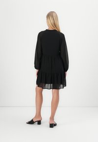 Crna haljina s prozirnim dugim rukavima, skupljenim strukom i stepenastim volanima; uparen s crnim sandalama na blok petu. Jednostavan i elegantan dizajn.