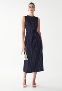 Marineblauwe mouwloze jurk met witte verticale strepen, geknoopte taille-detail, midi-lengte. Model houdt een kleine witte handtas vast en draagt sleehakken.