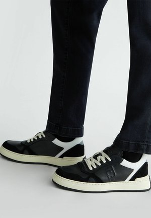 Sneaker in pelle bianca e nera con tomaia texturizzata, suola in gomma crema e lacci a contrasto. Branding visibile sul lato.