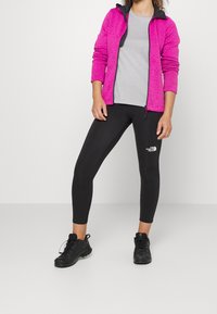 Giacca rosa con zip foderata in pile, t-shirt grigia, leggings neri con logo e scarpe da ginnastica nere. Presenta una vestibilità slim e un tessuto testurizzato.