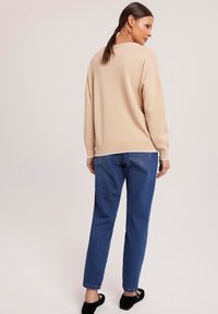 Beżowy sweter z dzianiny z opadającymi ramionami, w zestawie z niebieskimi dżinsami. Stylizacja charakteryzuje się luźnym krojem i delikatnym prążkowanym wykończeniem u dołu.