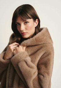 Kobieta z prostymi brązowymi włosami, ubrana w puszysty, jasnobrązowy futrzany płaszcz, trzymająca kołnierz blisko szyi na neutralnym tle.