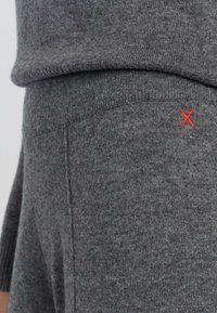 Grauer Strickpullover mit geripptem Kragen und Bündchen, sichtbarer Naht und einem kleinen roten gestickten X-Detail an der Seite.