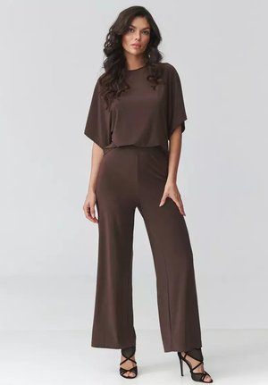 Kobieta o długich kręconych włosach, ubraną w luźną brązową bluzkę z krótkim rękawem i szerokie spodnie, stojąca w czarnych, paskowych szpilkach na jednolitym tle.