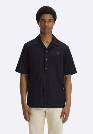 Giovane uomo in piedi, indossa una camicia nera a maniche corte con bottoni e un piccolo stemma, e pantaloni beige su uno sfondo semplice.