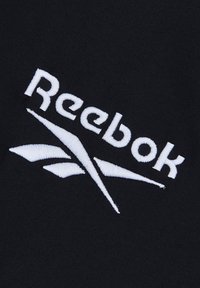 Logo Reebok przedstawia biały haftowany tekst na czarnym tle materiału, z stylizowanym geometrycznym wzorem pod nazwą marki.